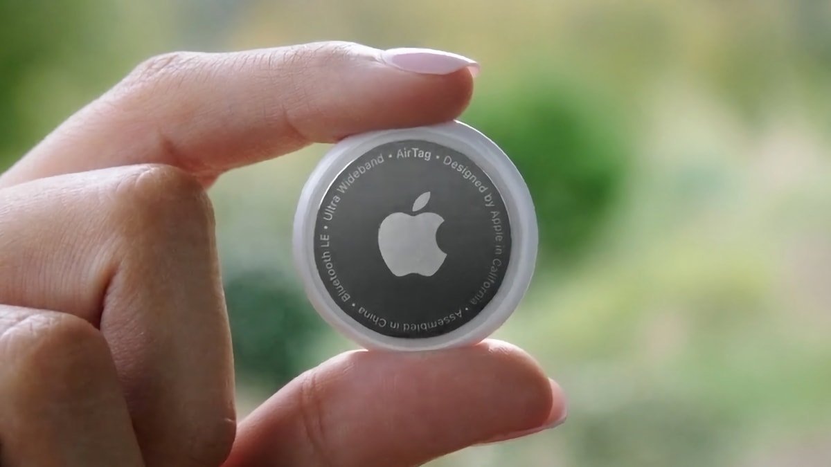 Hand holding a small round Apple AirTag tracker between thumb and finger, silver with Apple logo centered, blurred green outdoor background behind it