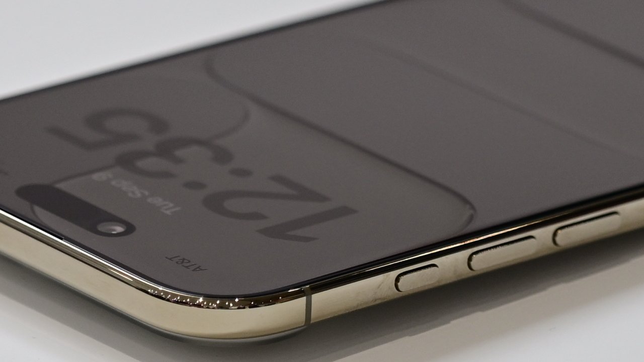 Close-up of a modern smartphone lying flat, showing reflective dark screen with faint clock numbers, rounded gold metal frame, and side volume and power buttons