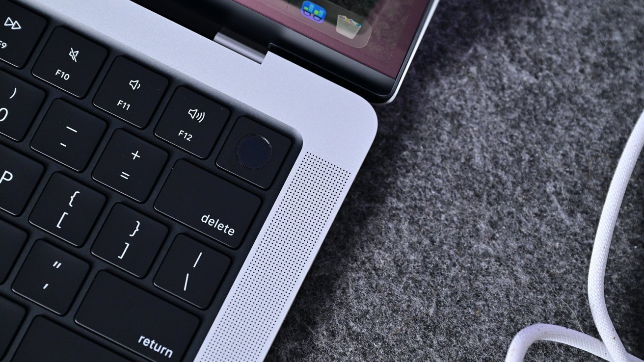 Close-up of a laptop keyboard corner with delete key, Touch ID button, speaker grille, open screen edge, and a white charging cable on a gray felt surface
