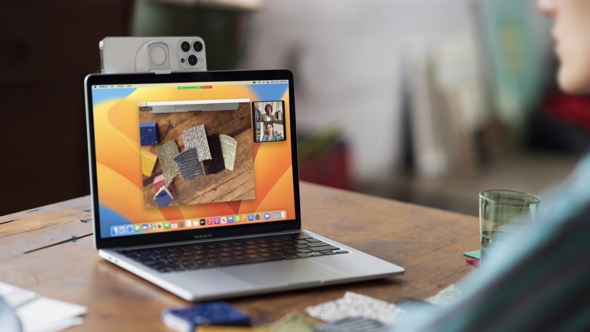 Open MacBook Pro on a wooden desk showing a video call and fabric samples, with an iPhone mounted on top as a webcam and a glass nearby.