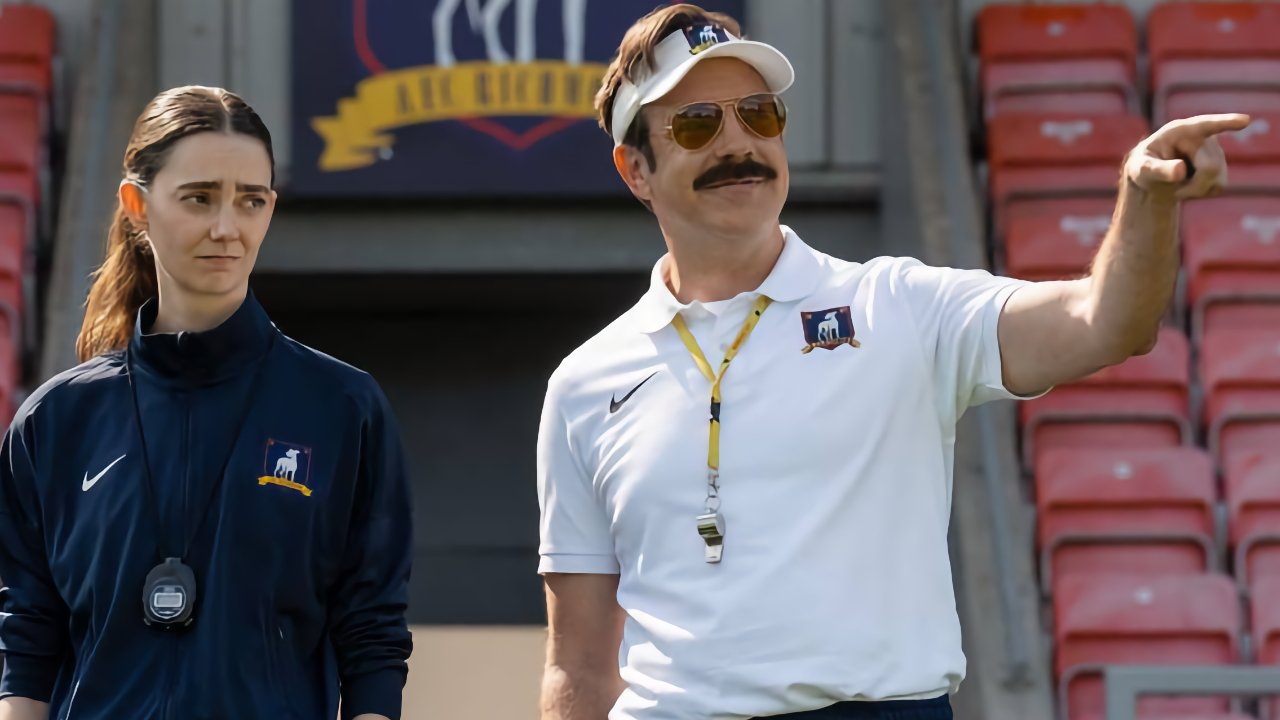 Soccer coaches on a stadium sideline, one in a white visor smiling and pointing, the other in a navy tracksuit watching with a serious expression, red seats in background