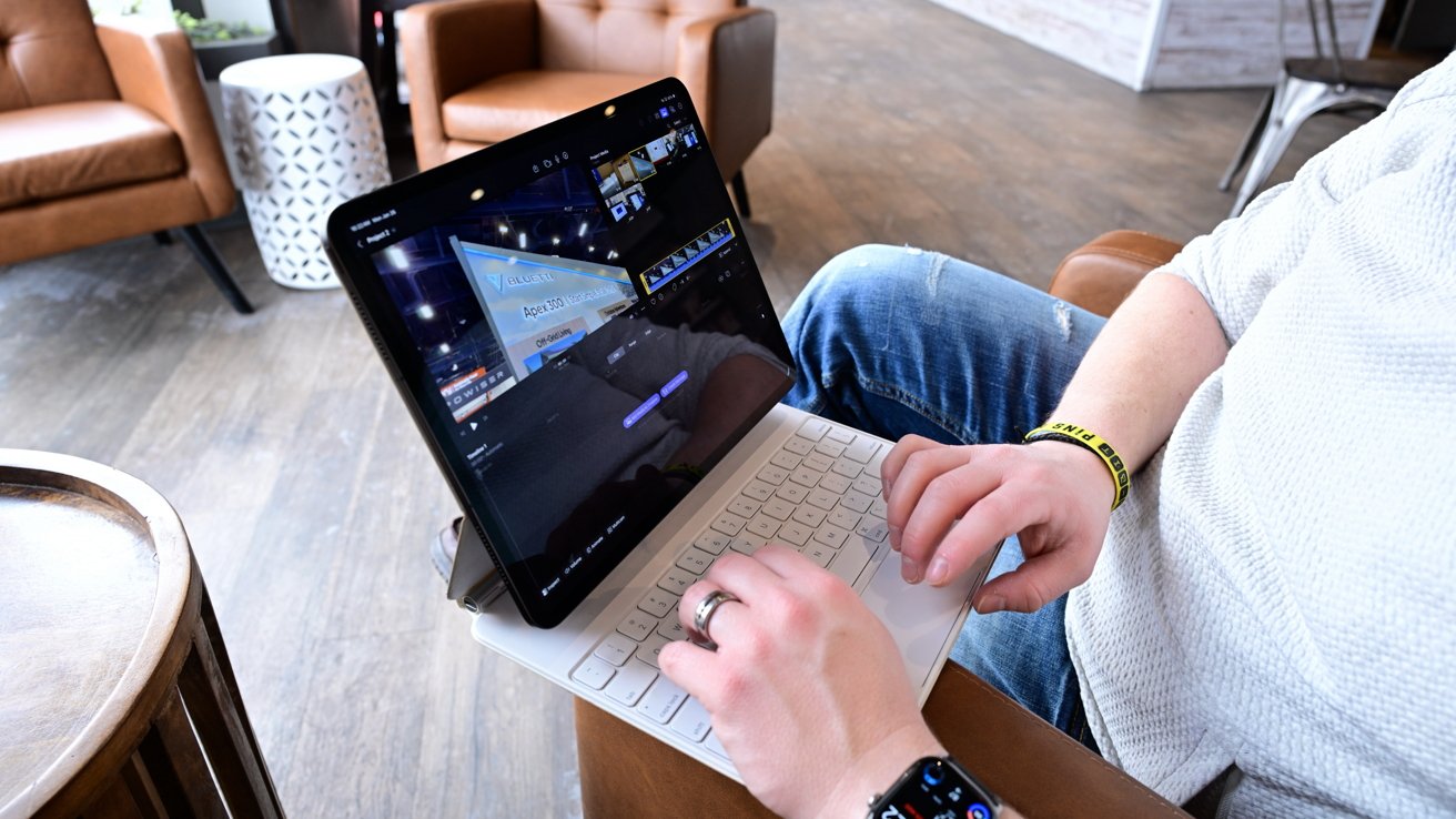 Person sitting in a modern lounge, typing on a tablet with detachable keyboard, editing video on screen, wearing jeans, a smartwatch, and a yellow wristband near leather chairs and tables
