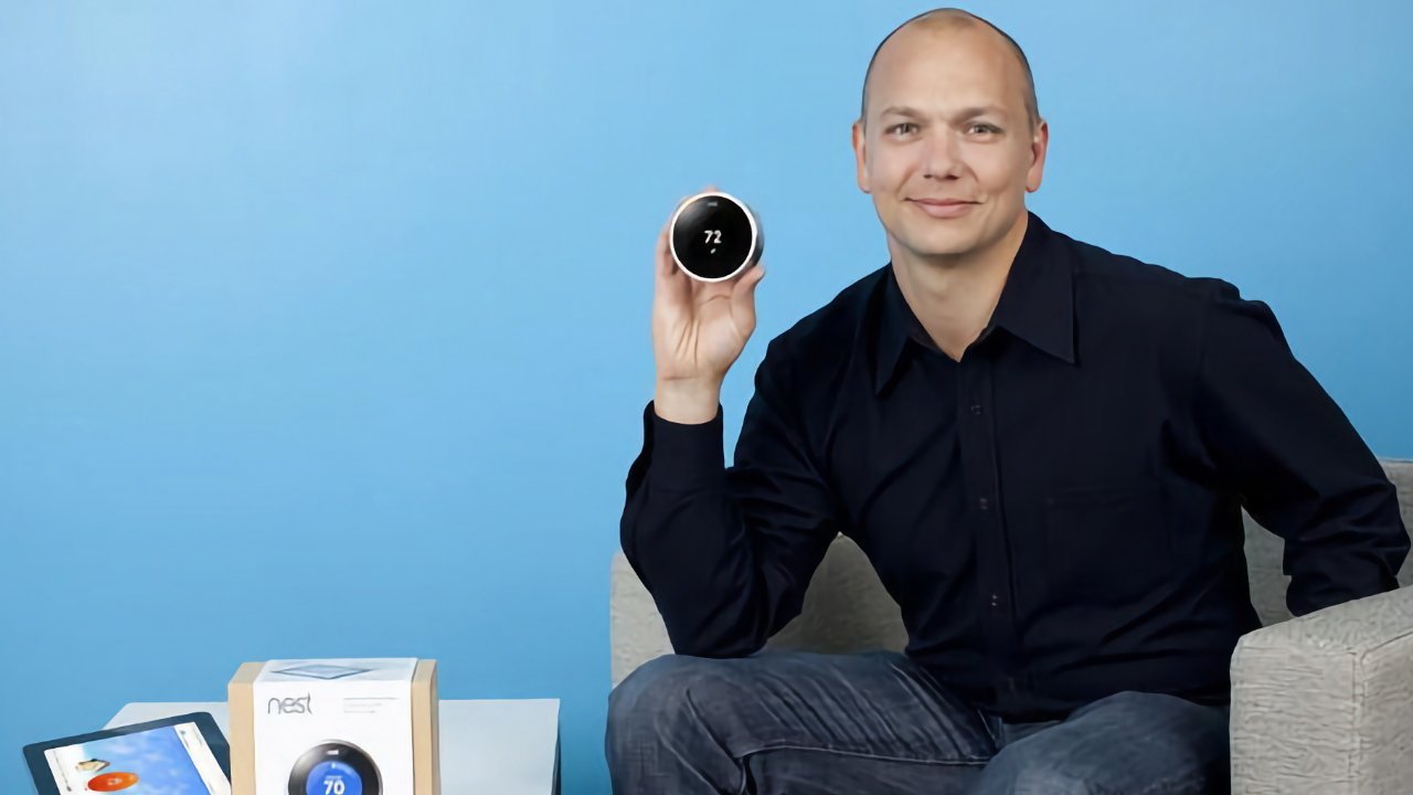 Smiling man seated against a blue wall, holding up a round digital thermostat displaying 72 degrees, with product packaging and a tablet on a table beside him