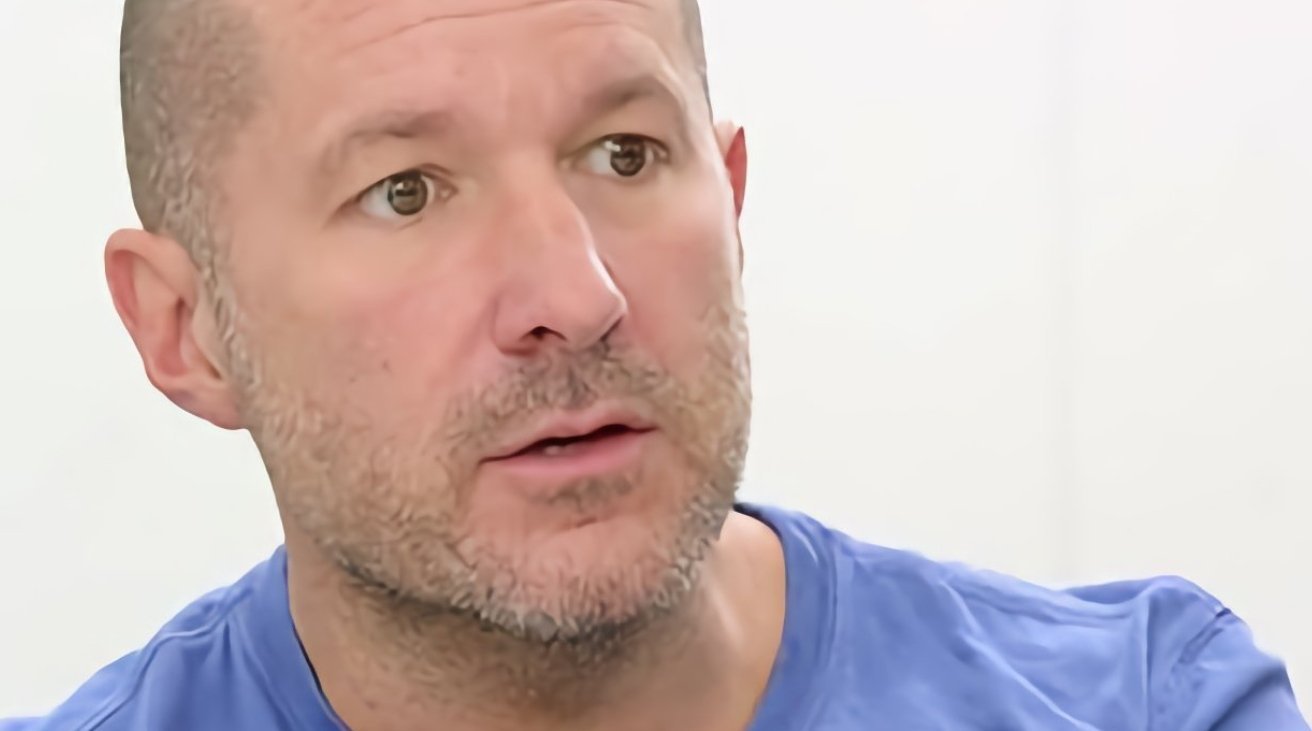 Bald middle-aged man with stubble wearing a light blue shirt, looking slightly to the side with a thoughtful, attentive expression against a plain white background