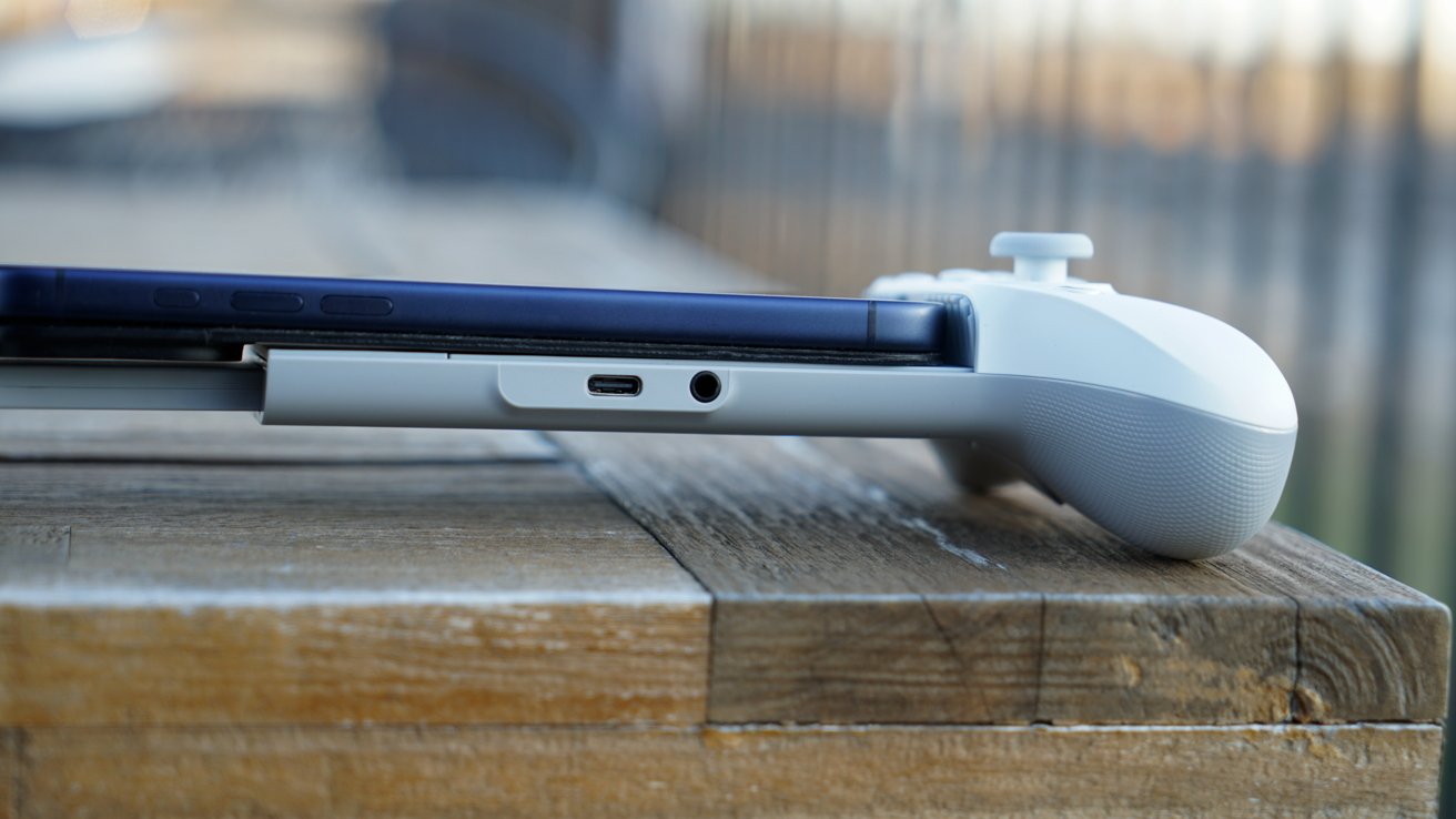 Side view of smartphone docked in white game controller grip on wooden table, showing USB-C ports and headphones with blurred outdoor background