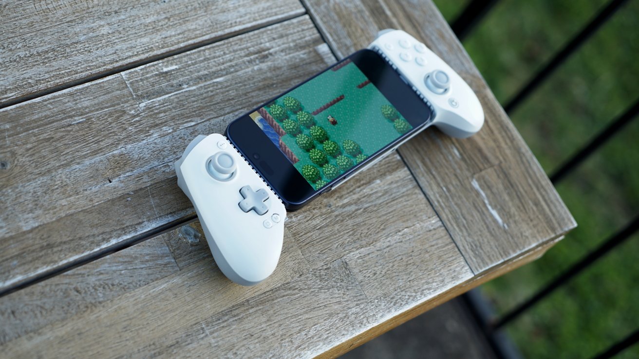 A smartphone clipped to a white game controller, showing a colorful pixelated adventure game, resting diagonally on a weathered wooden outdoor table with grass blurred in the background
