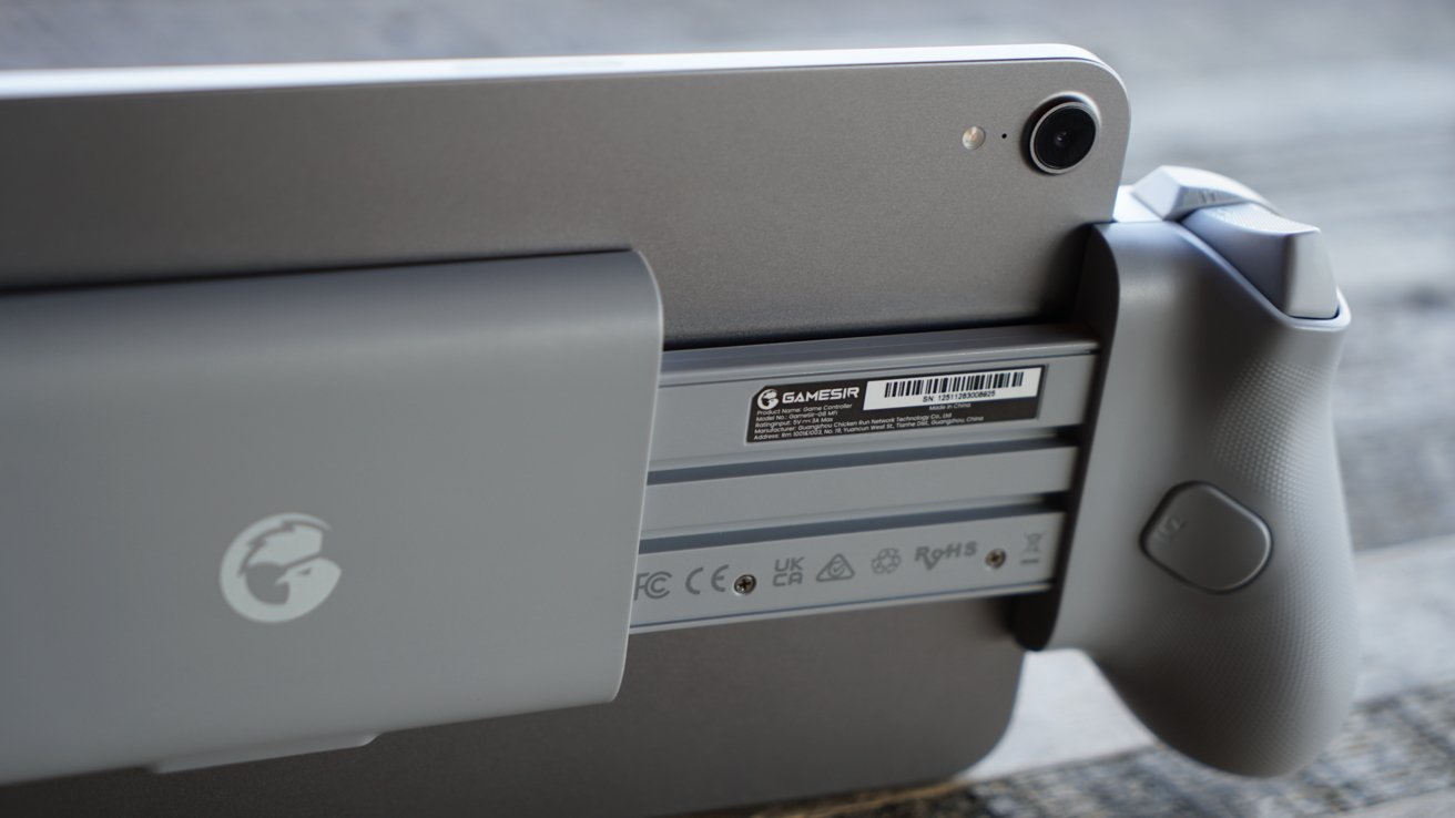 Close-up of a tablet attached to the gray handle of a GameSir game controller, showing the rear camera, textured handle, product label and certification logos on a wooden surface