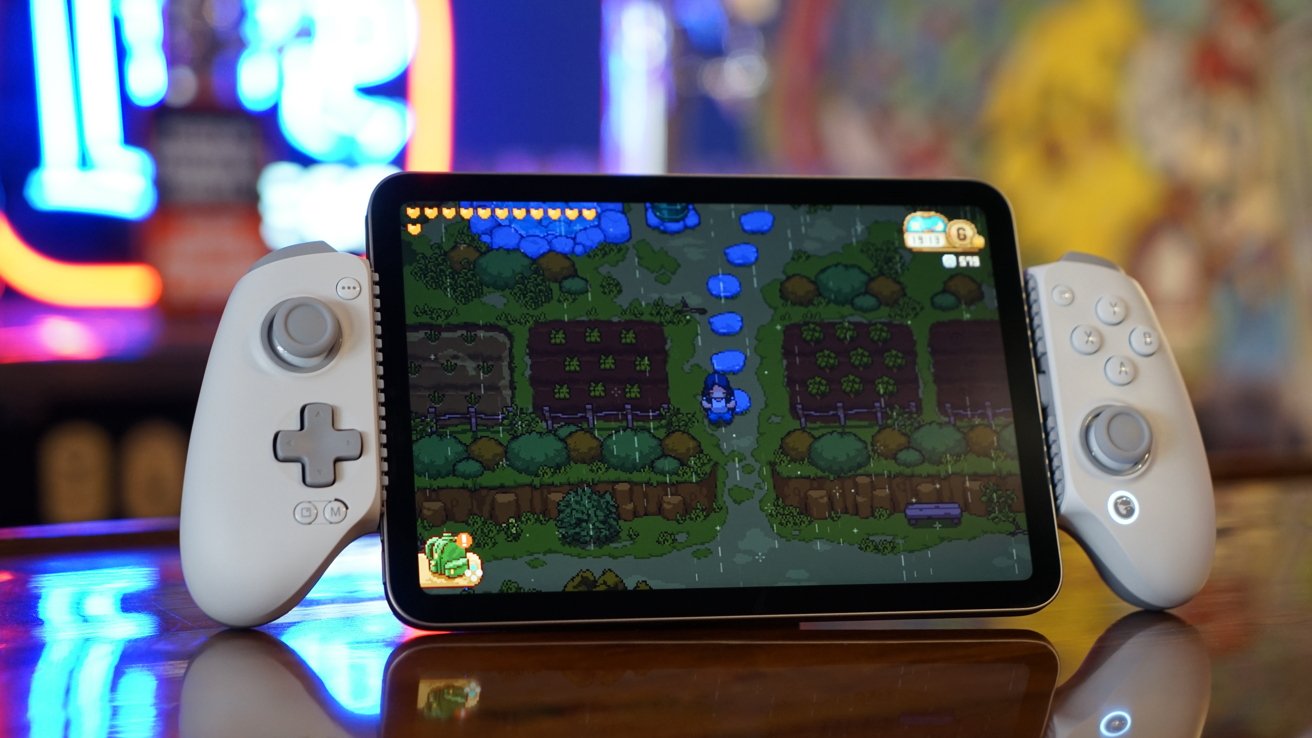 An iPad mini showing a pixelated farm game while in a white game controller grip with neon and wall art blurred in the background.