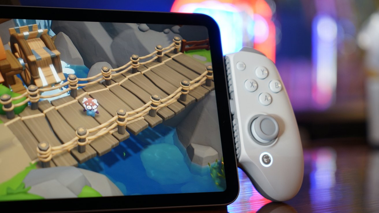 A tablet showing a colorful cartoon game with a small character on a wooden bridge over water, next to a white handheld game controller on a reflective surface.