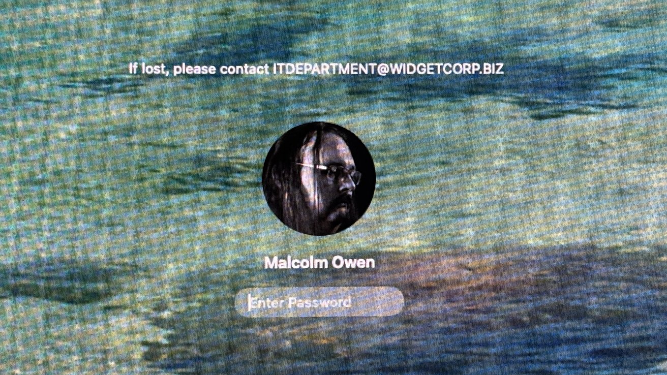 Computer login screen showing circular user photo of a man with glasses and long hair, username Malcolm Owen, password field, and lost-device contact email over a blurred water background