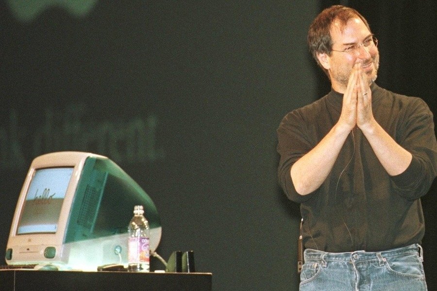 Man in black turtleneck and jeans smiling on stage, hands clasped, standing beside a colorful translucent desktop computer and water bottle against a dark background