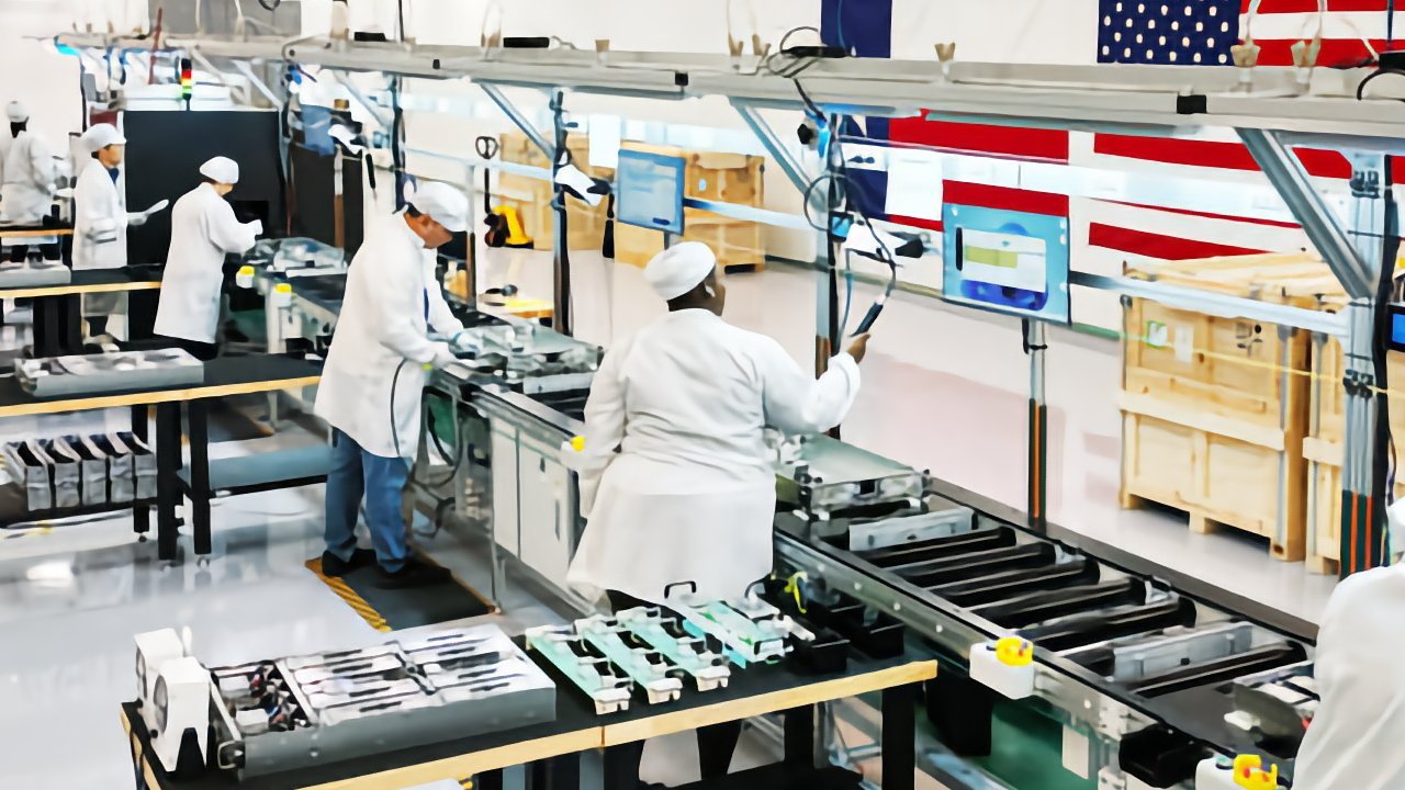 Workers in white lab coats and caps operate equipment along a clean manufacturing assembly line, with tools, conveyor belts, computer monitors, and large American flags hanging in the background