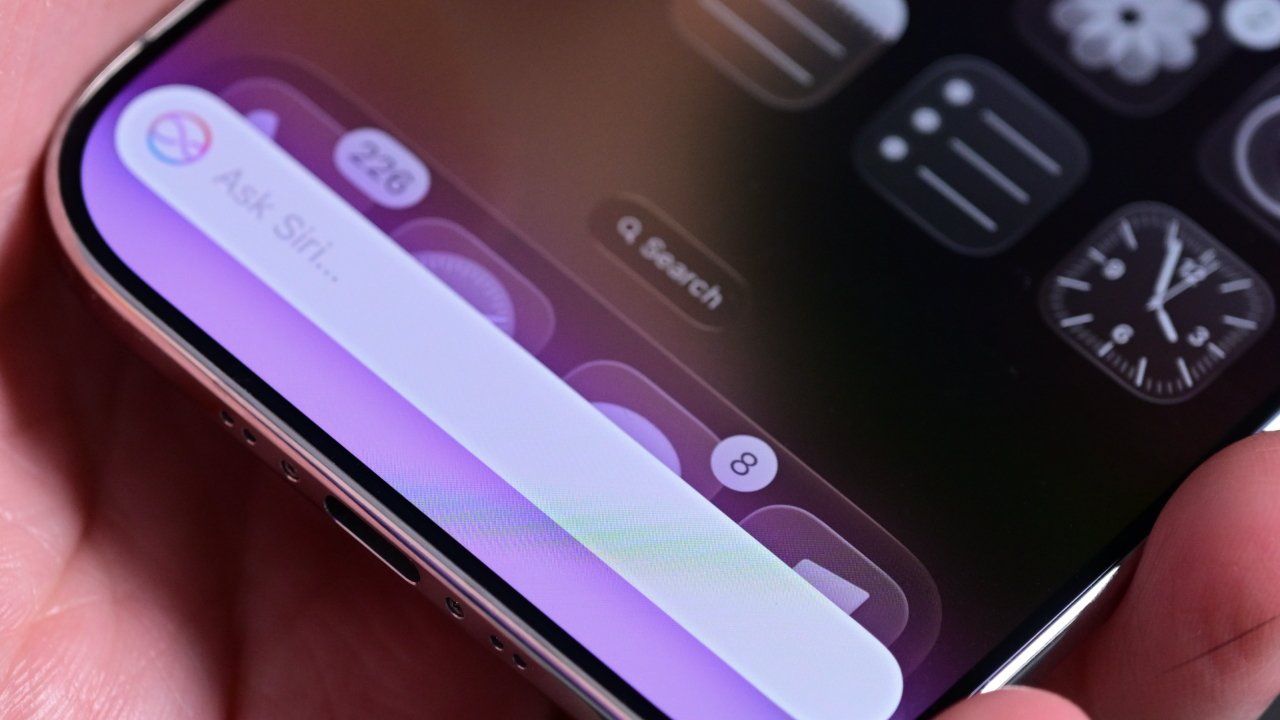 Close-up of an iPhone screen showing the Ask Siri text field and app dock, held in a hand, with blurred home screen icons and a clock widget in the background