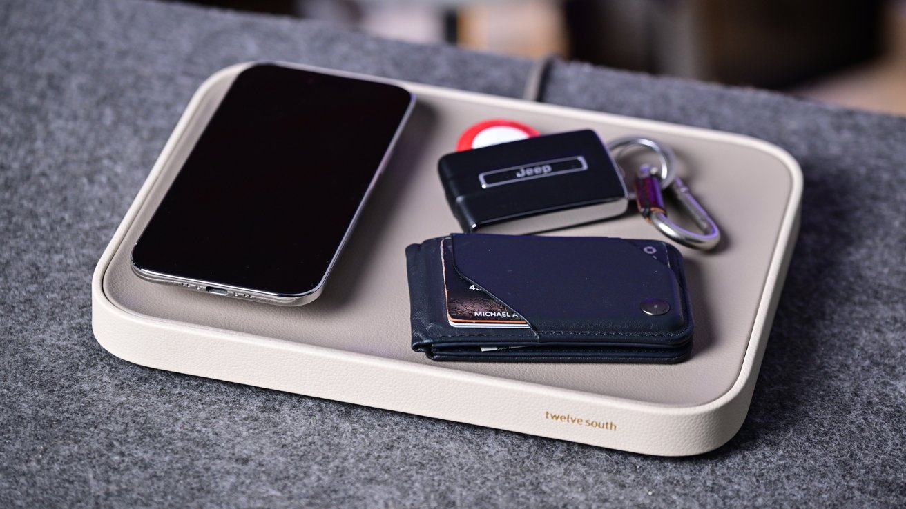 Beige table tray with smartphone, slim card wallet, car key fob, metal key ring and small round tracker resting on gray fabric surface