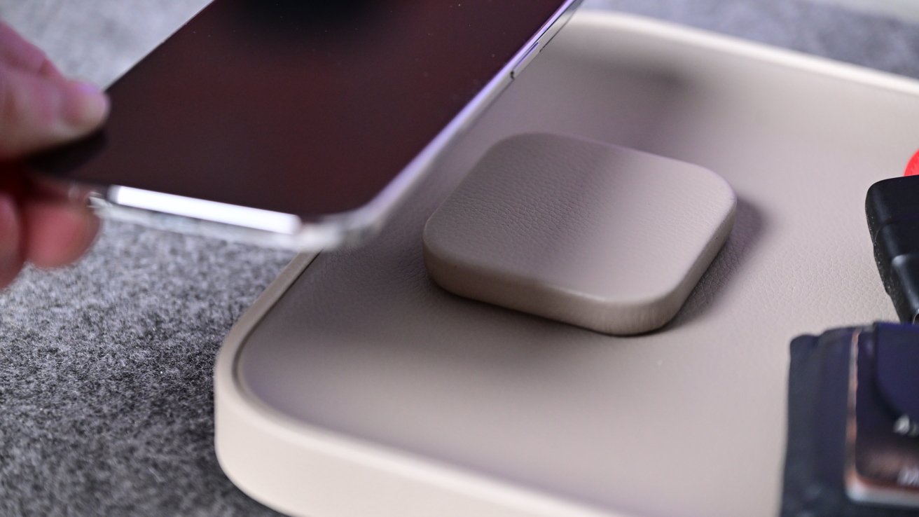 Hand holding smartphone over small square wireless charging pad on tray with keys and wallet nearby on textured surface