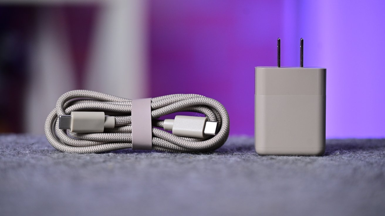A curled gray USB-C charging cable next to a matching rectangular wall power adapter, both resting on a soft gray surface with a blurred purple background
