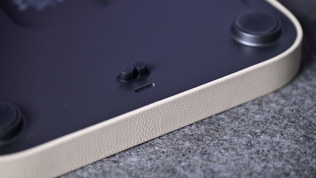 Close-up of rounded corner of device with textured beige edge, dark underside with two rubber feet, small power switch and narrow slot, resting on gray cloth surface