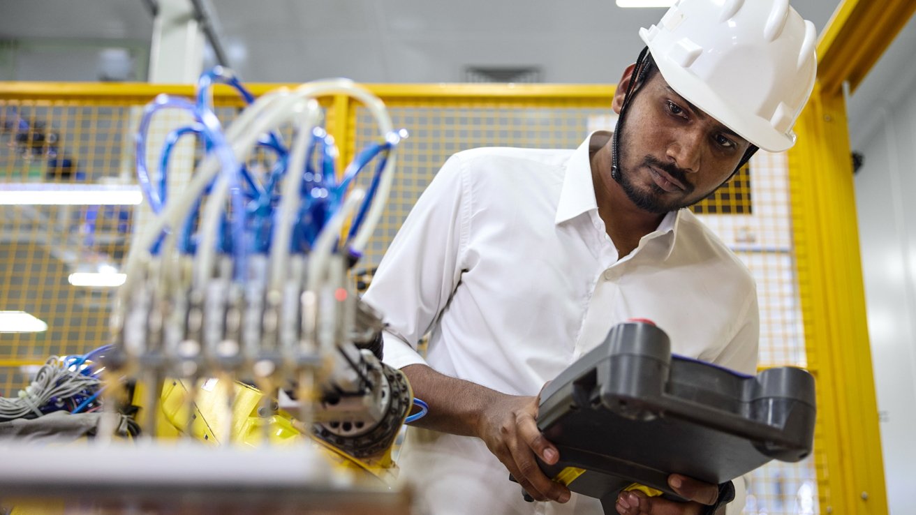 Engineer in white hard hat closely inspecting industrial machinery, holding a handheld control device, surrounded by cables and equipment inside a modern manufacturing or automation facility