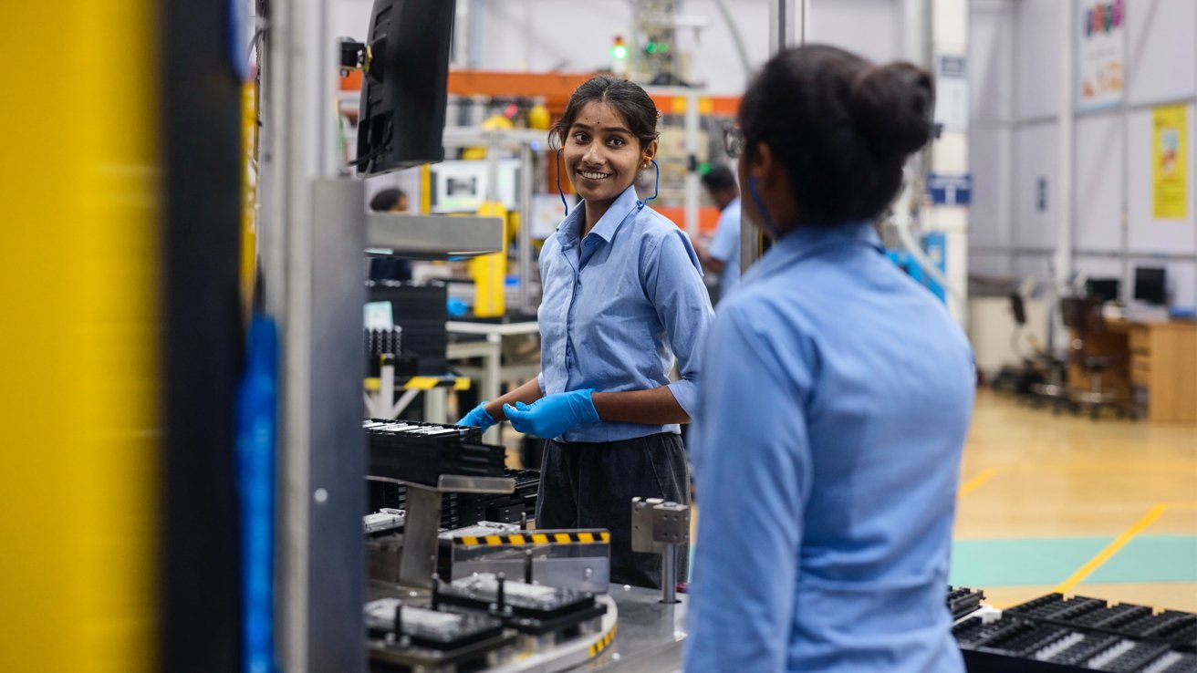 Two women in blue shirts and gloves work at a factory assembly line, smiling and talking amid industrial equipment, trays of parts, and a spacious, brightly lit workspace.