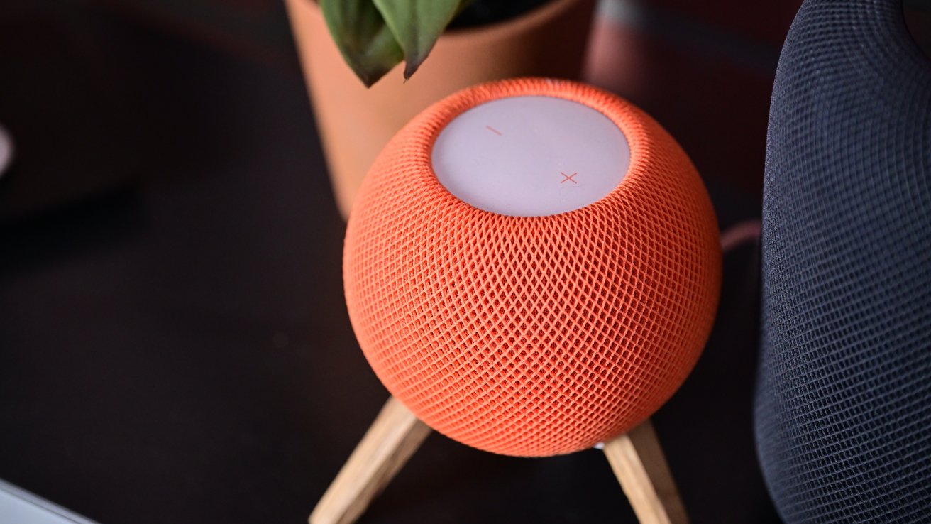 An orange HomePod mini by a gray HomePod on a table next to a potted plant