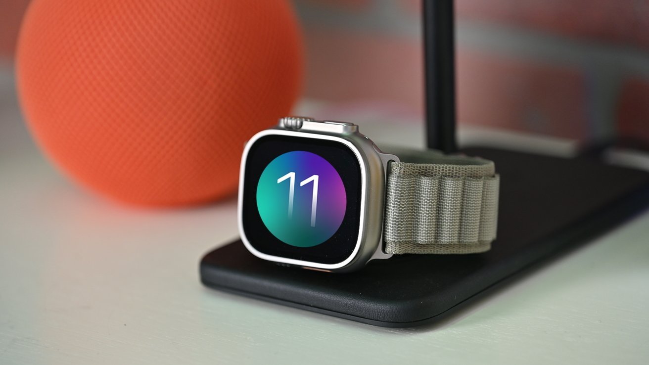 Silver smartwatch with fabric band resting on a black charging pad, screen showing a colorful circle with number 11, with an orange textured speaker blurred in the background