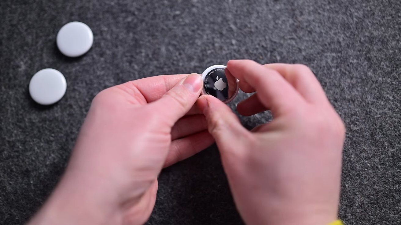 Hands holding a shiny Apple AirTag tracker above a gray fabric surface, with two other white circular AirTags lying nearby