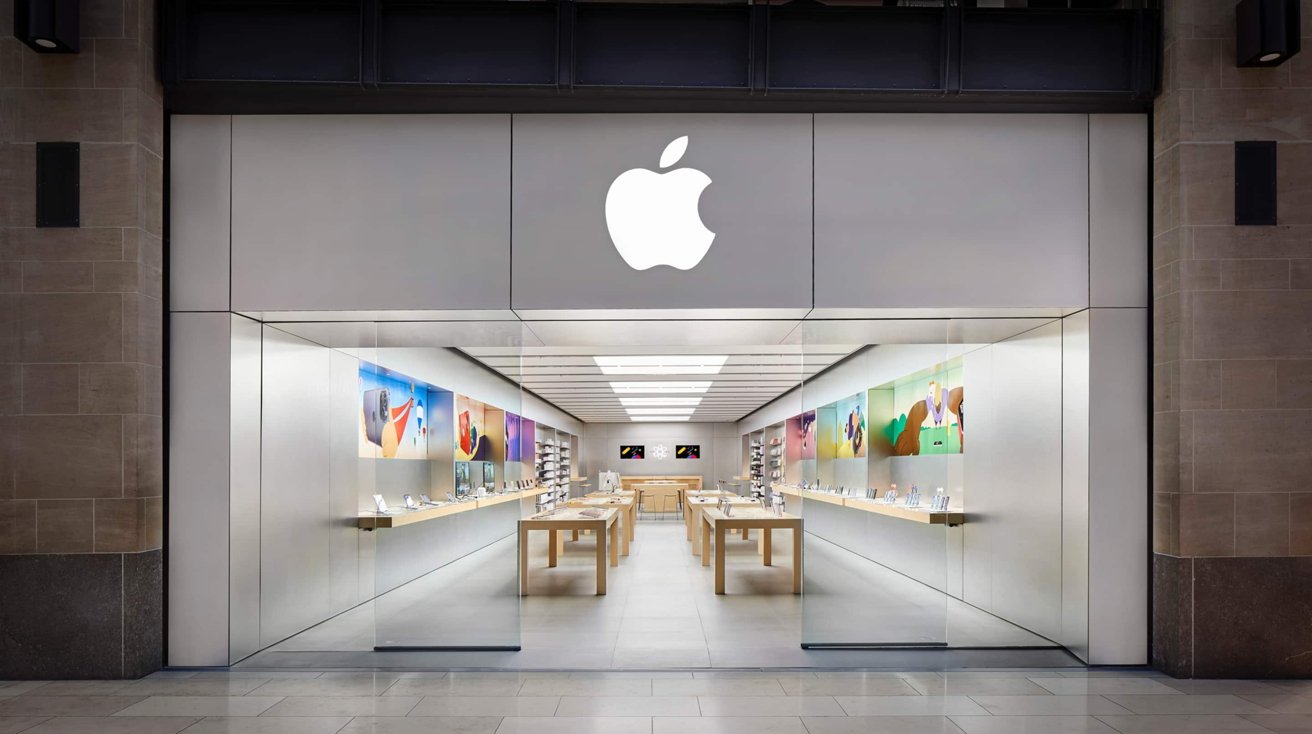 Bright, modern Apple retail store with large glowing Apple logo, glass entrance, wooden display tables, and colorful product posters lining both walls inside a well-lit shopping mall corridor