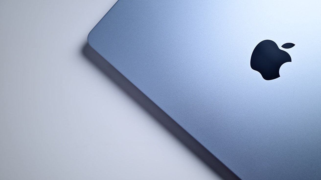 Close-up of a blue-gray Apple laptop lid on a light background, showing the black Apple logo near the right edge and smooth metallic surface