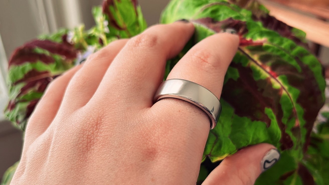 Hand wearing a wide silver ring on the middle finger, held against large green and burgundy leaves, with a thumb featuring a small abstract black-and-white nail design
