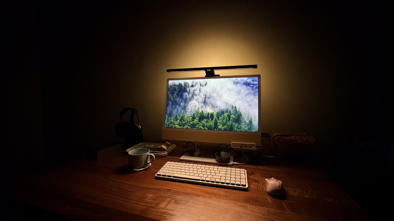Cozy dimly lit wooden desk with computer displaying misty forest, slim light bar above monitor, compact keyboard, white mouse, headphones, coffee mug, and small accessories nearby
