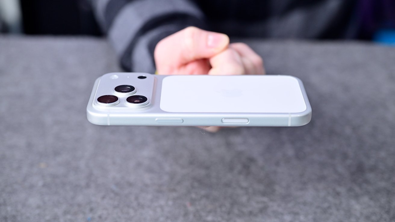 Hand holding a white smartphone horizontally above a gray surface, showcasing the side buttons and raised triple rear camera module on the back