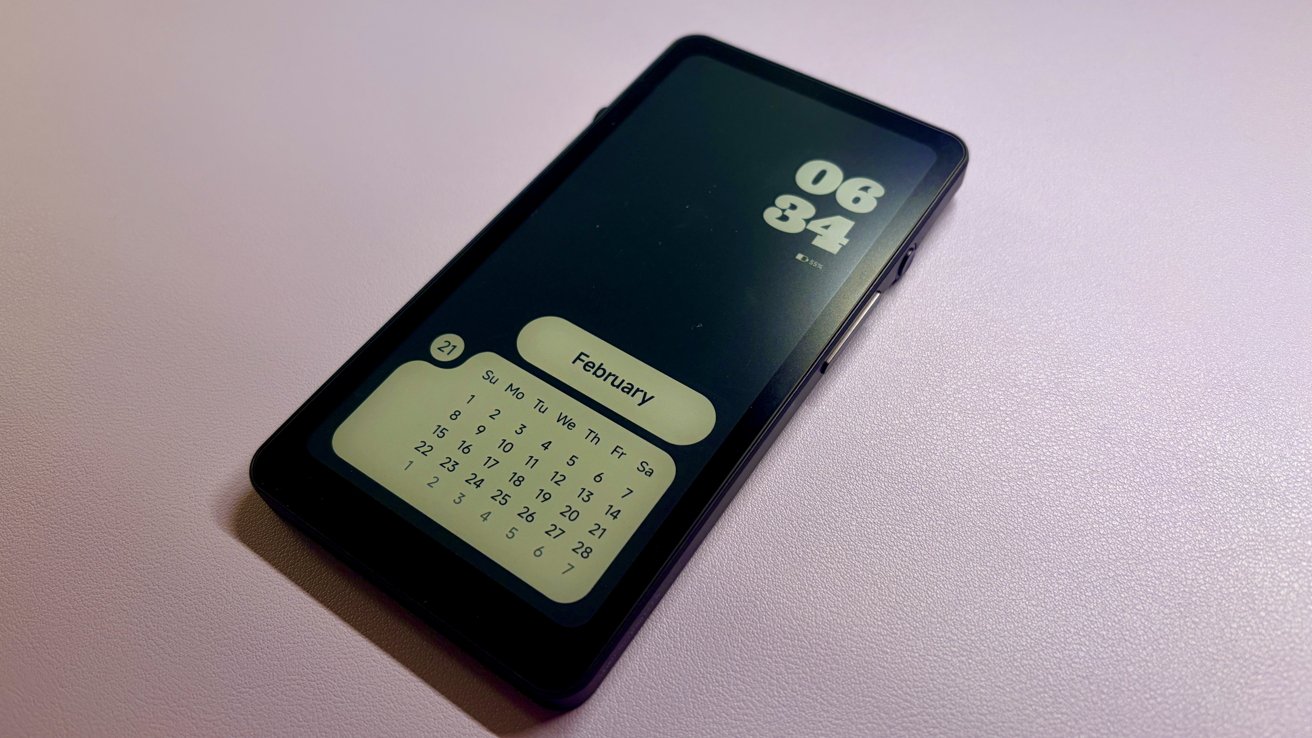 Black smartphone on a light pink surface, screen showing large digital clock, February calendar widget, and date 21, angled slightly with soft lighting and subtle shadow underneath