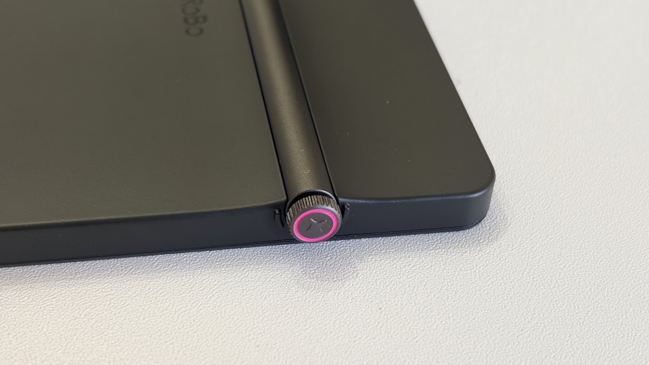 Крупный план тонкого черного устройства на слегка текстурированной поверхности: цилиндрический шарнир с небольшой металлической ручкой с розовым кольцом и изящным треугольным логотипом.