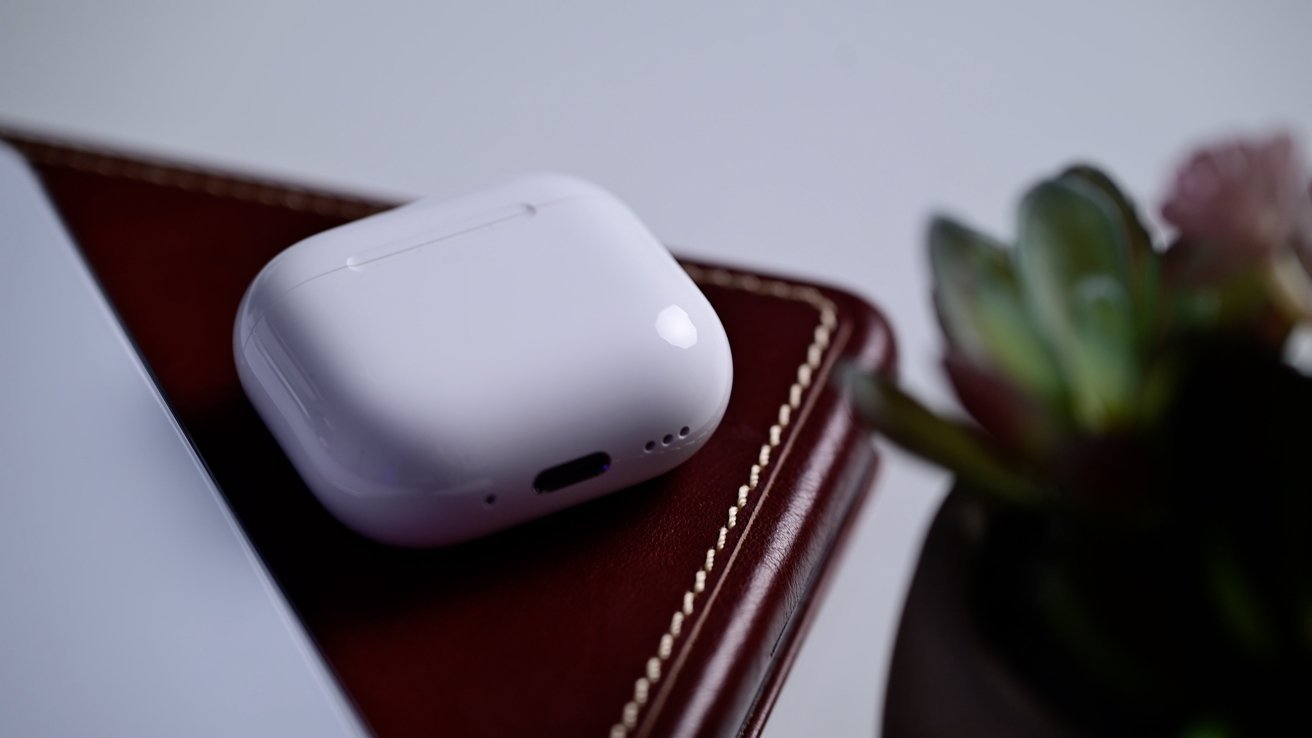 AirPods charging case resting on a brown leather laptop with blurred edges of the tablet on the left and a small succulent plant on the right