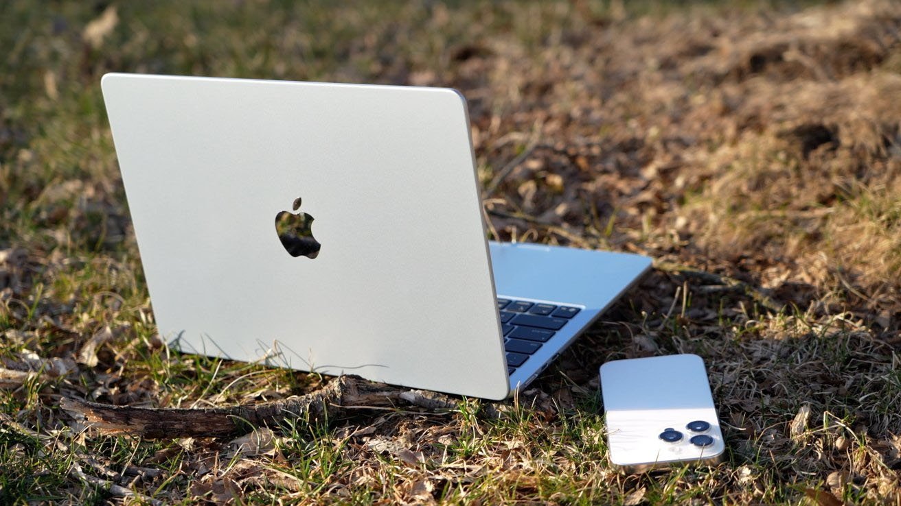 A silver Apple MacBook Air laptop open on dry grass next to a white smartphone, both resting outside in a sunlit, slightly dirty field with scattered twigs and patches of green.
