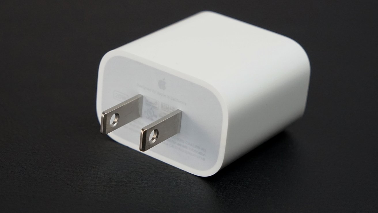 Apple wall power adapter with two flat metal pins, lying on its side on a dark surface, with minimal text and logo on the front of the plug
