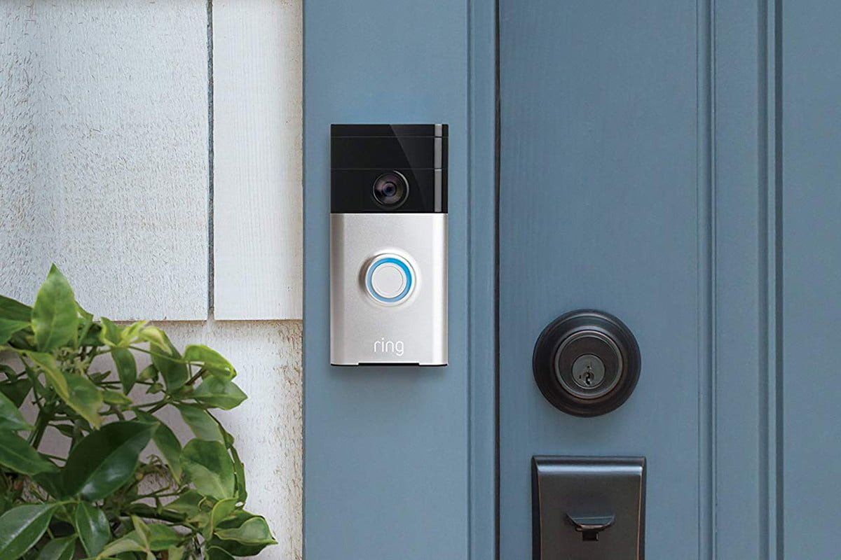 A smart video doorbell with a camera and a light-up button mounted next to a blue front door and lock, next to white cladding and a green leafy plant
