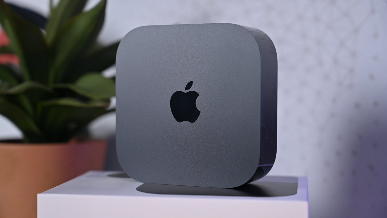 Space gray Apple Mac mini desktop computer standing vertically on a white surface, with a blurred green potted plant and soft patterned wall in the background