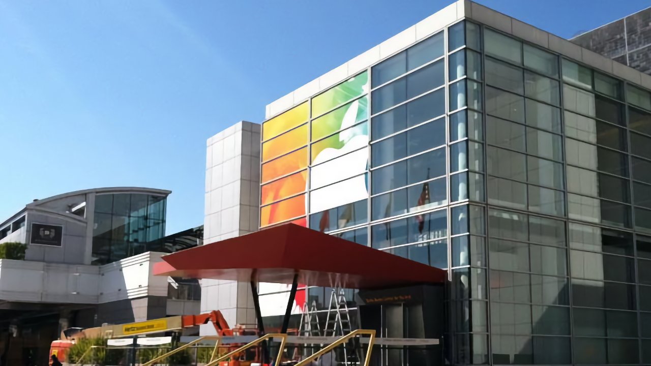 Modern glass convention center with large colorful Apple logo mural, red entrance canopy, and construction equipment outside under clear blue sky