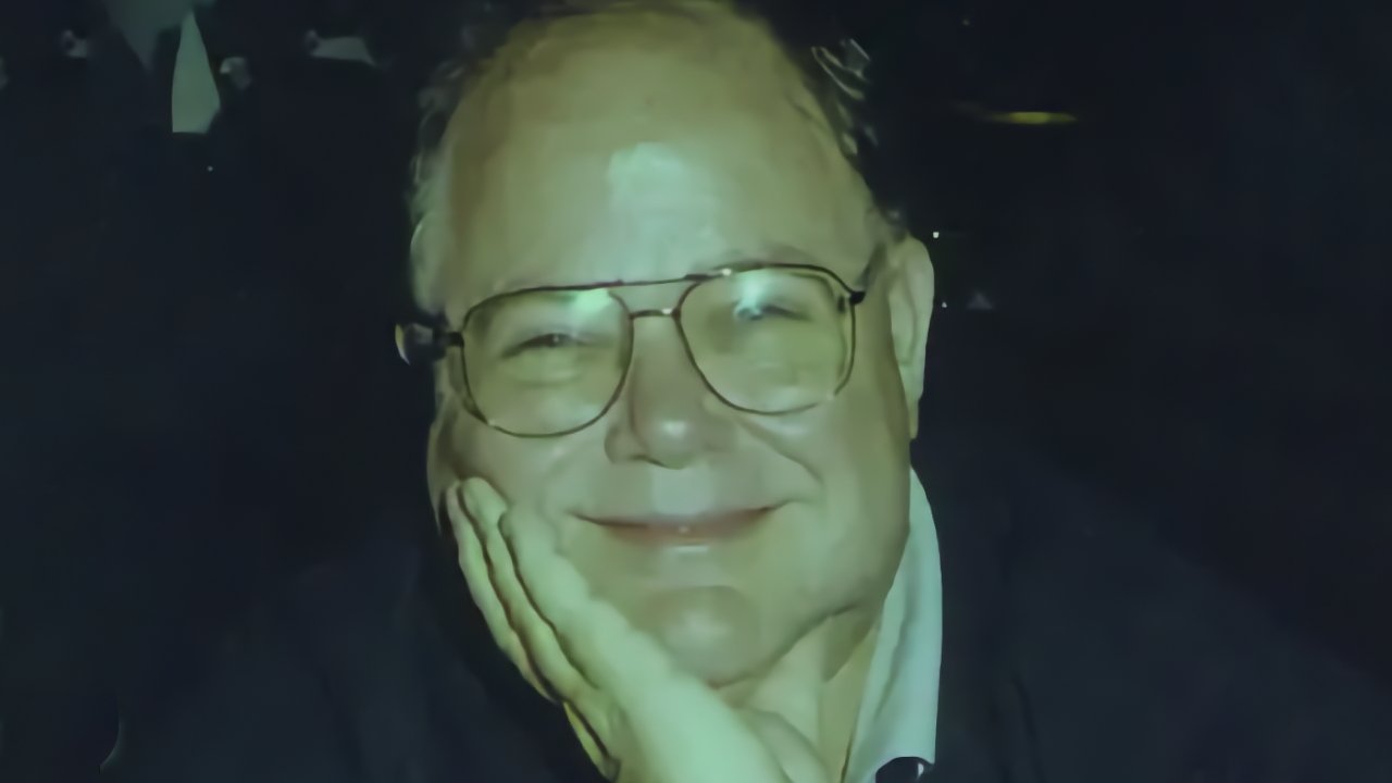 Smiling older man with glasses resting his chin on his hand, looking directly at the camera against a dark background