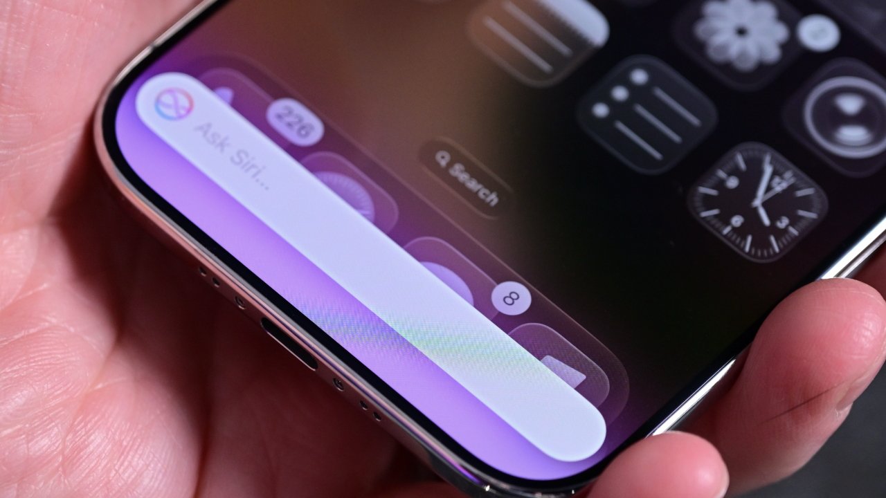 Close-up of a hand holding a smartphone showing a colorful Siri input bar on the home screen with app widgets, search field, and digital clock icons visible.