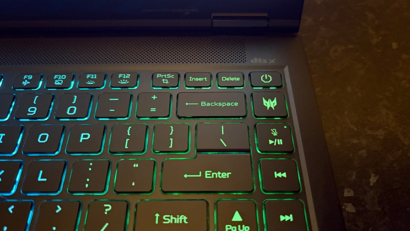 Close-up of a laptop keyboard with green backlit keys, showing Backspace, Enter, power button, media controls, and part of the speaker grill on a dark surface