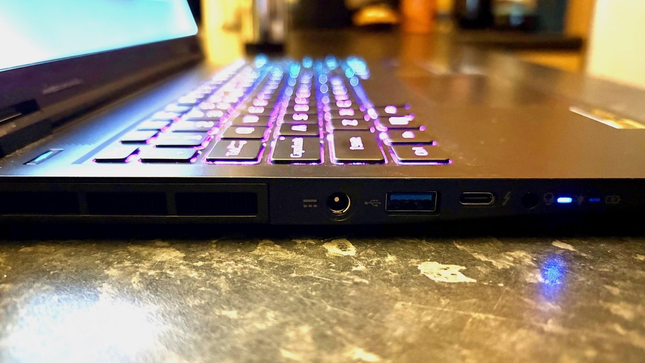 Side view of a slim laptop on a desk, showing glowing RGB backlit keyboard, various ports and vents along the edge, and small blue indicator lights near the front