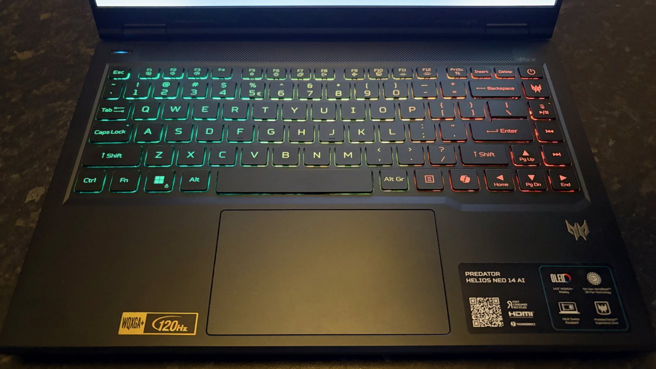 Open gaming laptop keyboard with RGB backlighting transitioning from green on the left to red on the right, large touchpad below, and branding stickers near the bottom right corner