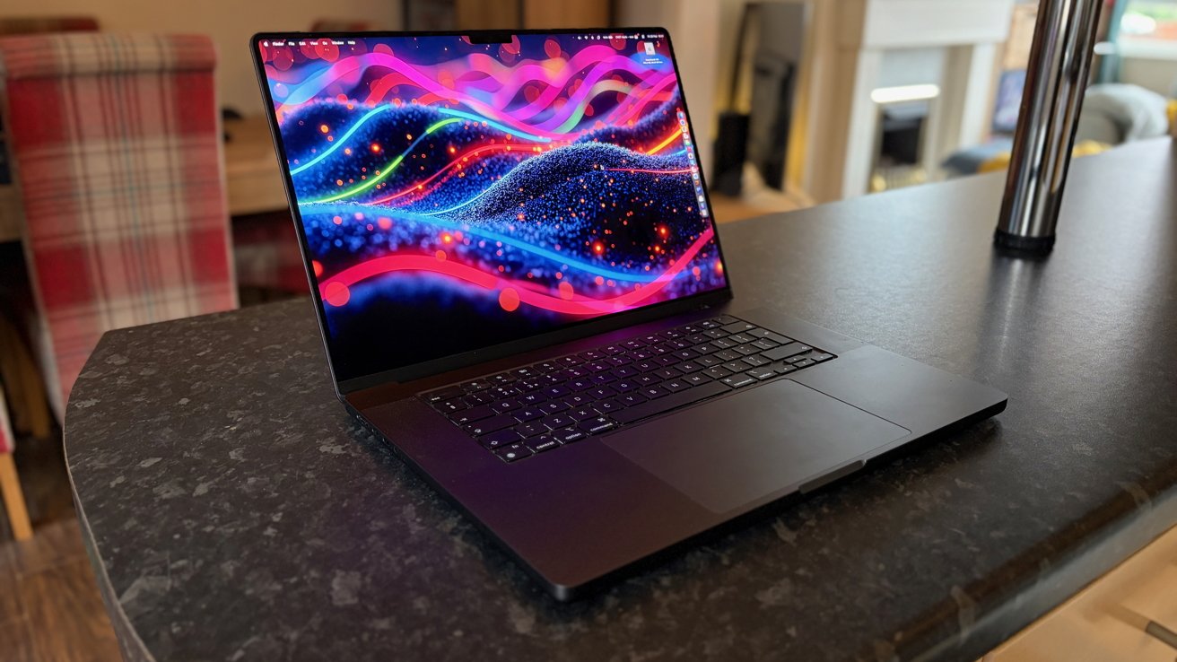 Open laptop on a dark kitchen countertop, displaying a vibrant abstract neon wallpaper with colorful waves and particles, in a softly lit modern home interior.