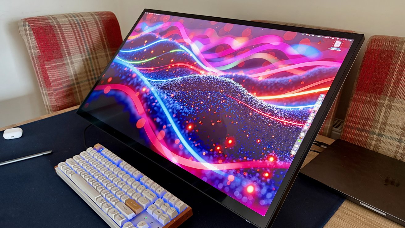 Large tilted monitor displaying colorful abstract waves on a desk, with a backlit mechanical keyboard, stylus, wireless mouse, and plaid-upholstered chairs in the background