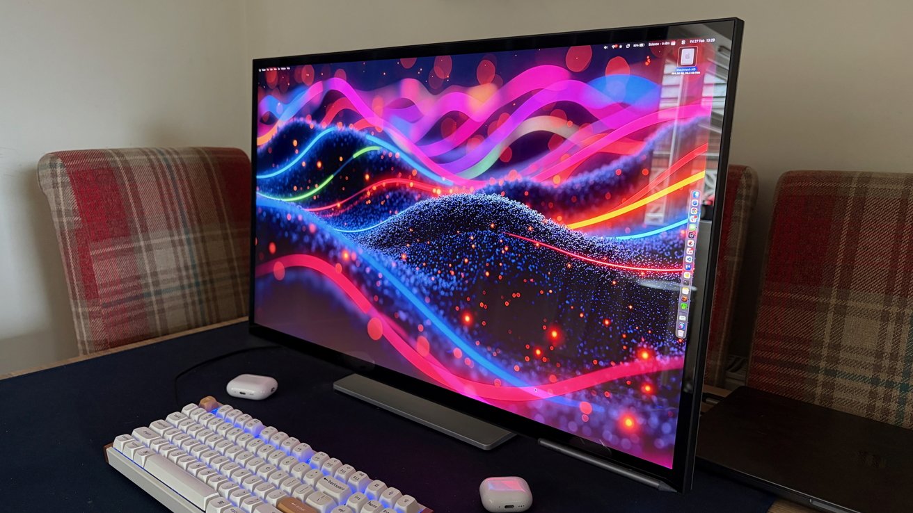 Large desktop monitor showing colorful abstract waves on a desk, with a mechanical keyboard, wireless earbuds case, and plaid-upholstered chairs in the background