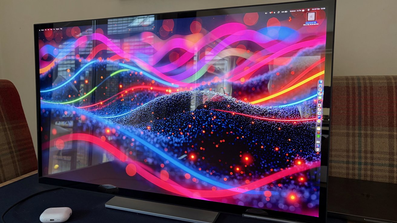 Large computer monitor on a desk showing colorful abstract waves and glowing dots, with Mac desktop icons visible along the right edge and a wireless earbud case in front