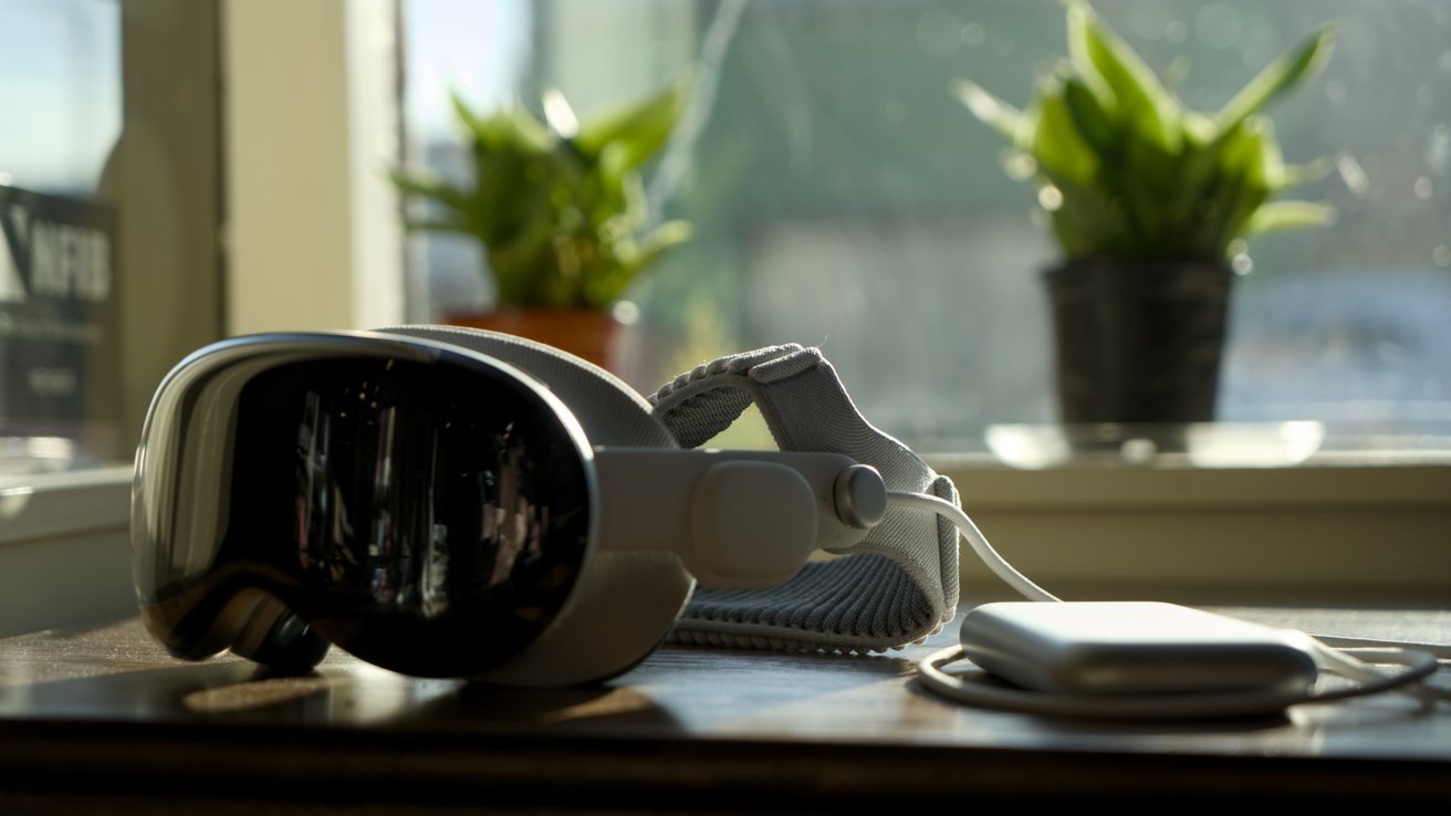 Sleek virtual reality headset and connected battery pack resting on a wooden table by a sunny window, with two small green potted plants blurred in the background