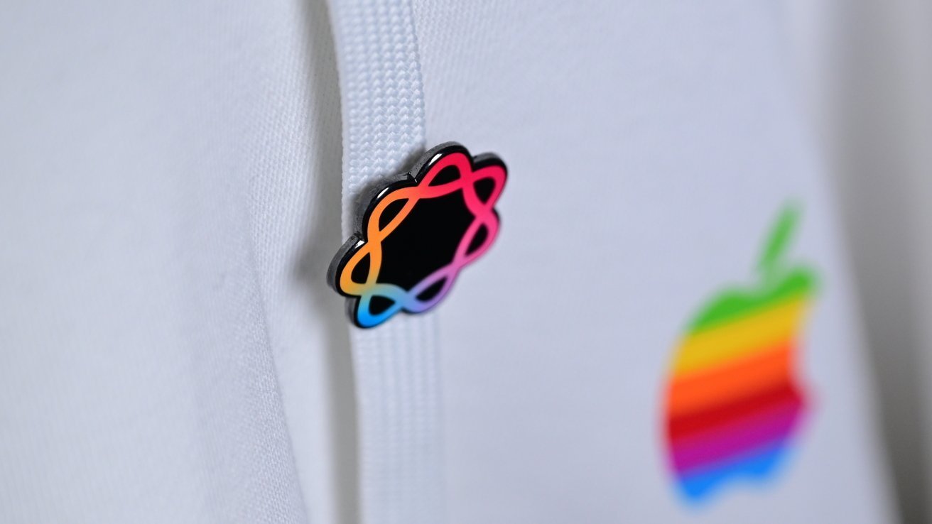 Close-up of a white fabric hoodie with a colorful gradient atom-shaped pin on the drawstring and a blurred rainbow-striped Apple logo in the background